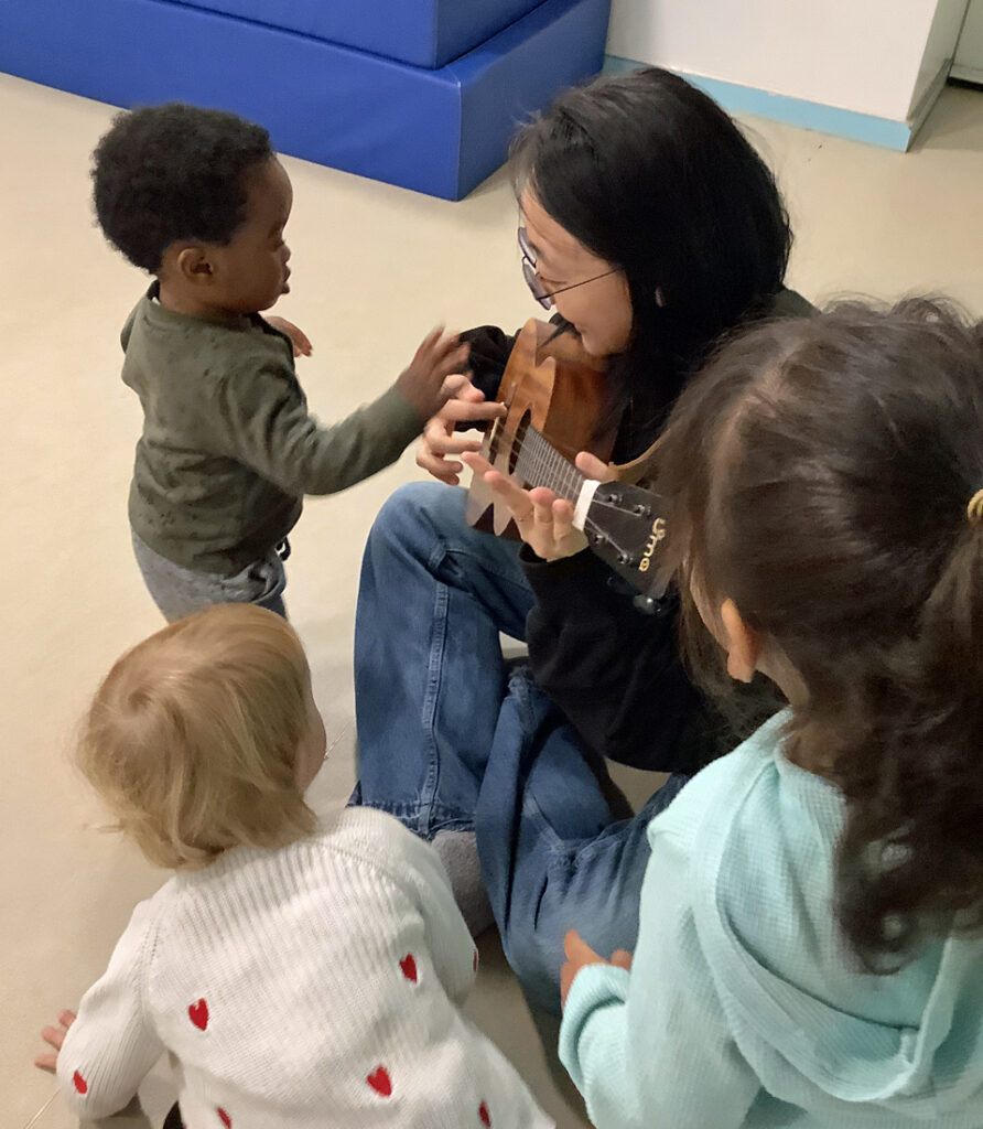 Eine Frau spielt auf einer kleinen Gitarre und Kita-Kinder schauen zu