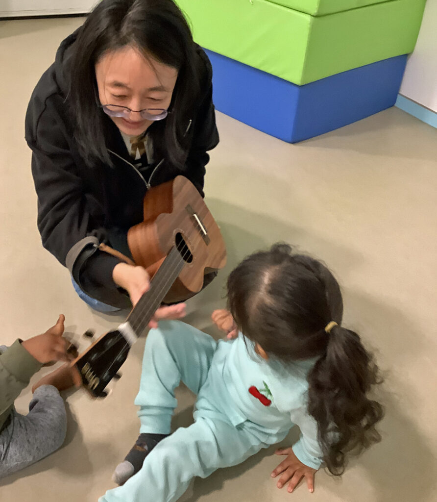 Eine Frau zeigt Kindern ihre kleine Gitarre und laesst sie das Instrument beruehren
