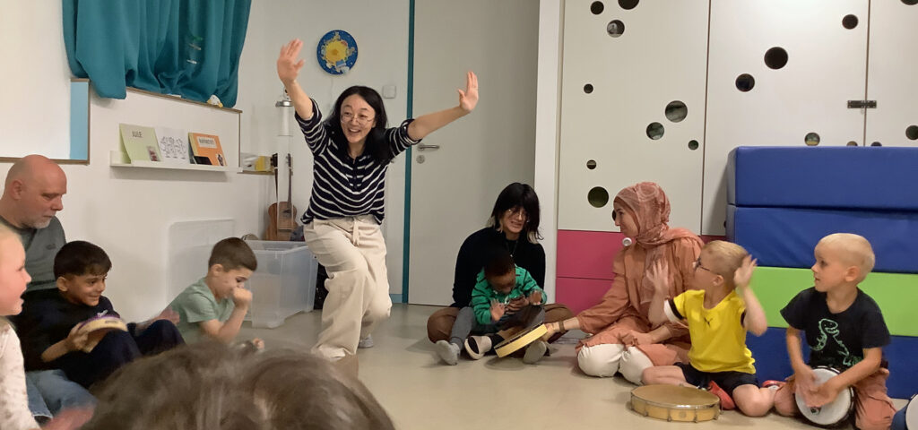 Eine Frau reckt froehlich die Arme in die Luft waehrend Kinder im Kreis sitzen und trommeln