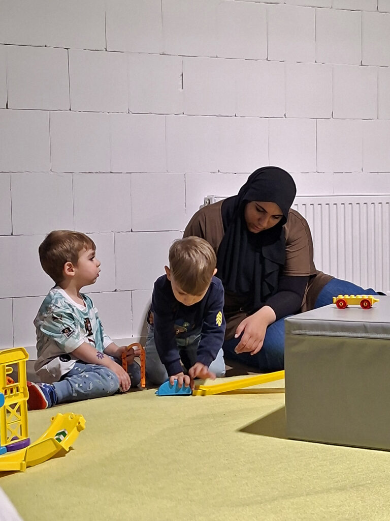 Eine Erzieherin sitzt mit Kindern auf dem Fussboden und sie bauen eine Bahn für Fahrzeuge