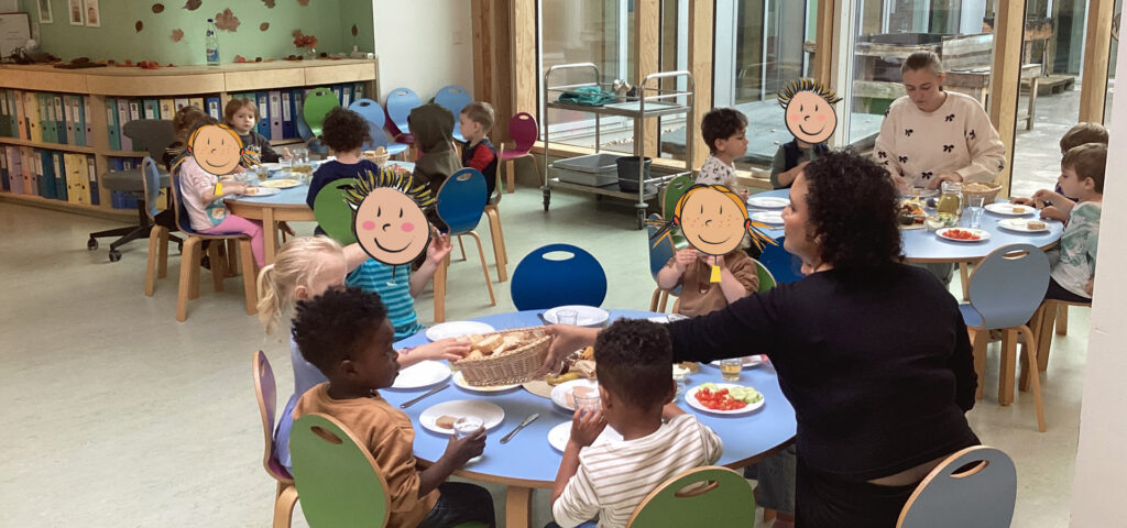 Kinder und Erzieherinnen sitzen an bunten Tischen in der Kita und essen