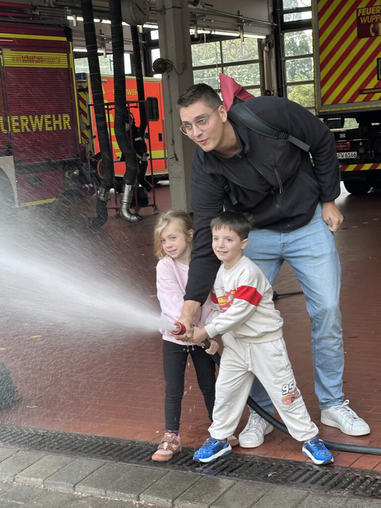 Zwei Kinder und ein Erzieher halten einen Feuerwehrschlauch und spritzen Wasser