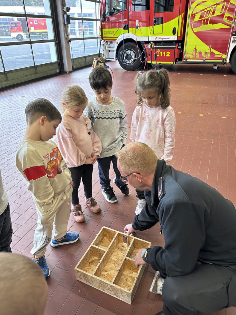 Ein Mitarbeiter der Feuerwehr erklaert den Kitakindern Fluchtwege anhand eines kleinen Haus-Modells