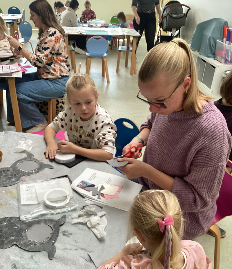 Muetter und Kinder sitzen an Tischen und schneiden Formen aus Pappe aus kleben mit einem Klebestift