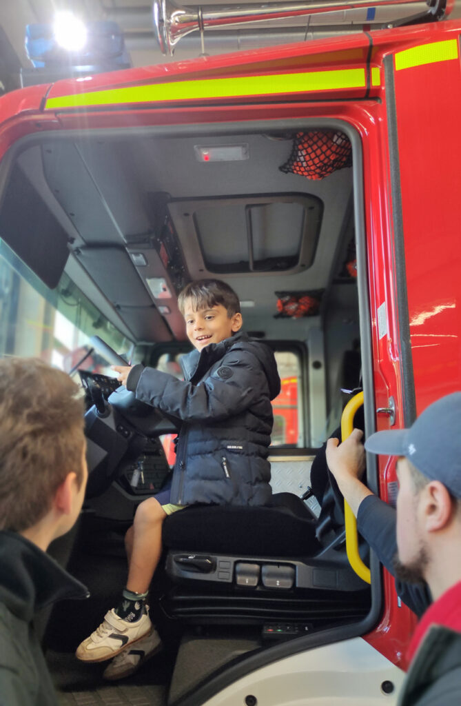 Ein Kind sitzt am Steuer eines grossen roten Feuerwehrautos und laechelt zwei Maennern zu